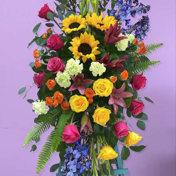 Tall standing floral spray with roses, sunflowers, lilies, and blue accents
