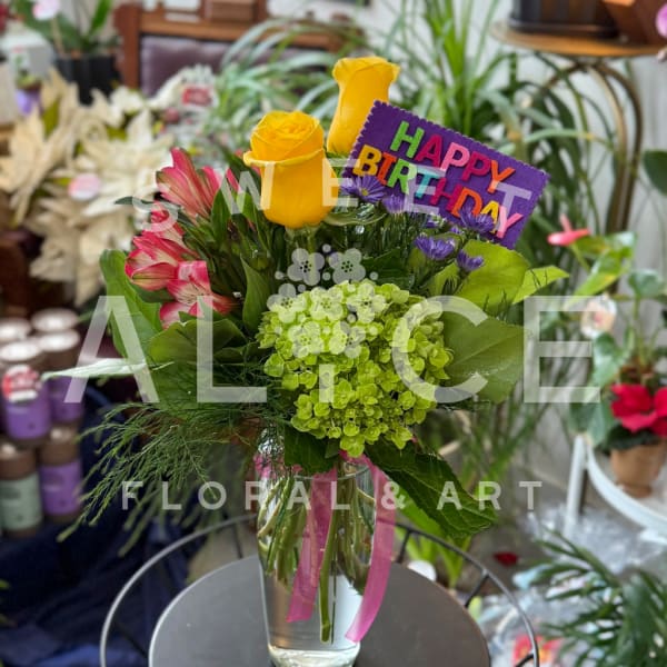 Bouquet of yellow roses, pink alstroemeria, and green hydrangea in a glass vase with a birthday sign.
