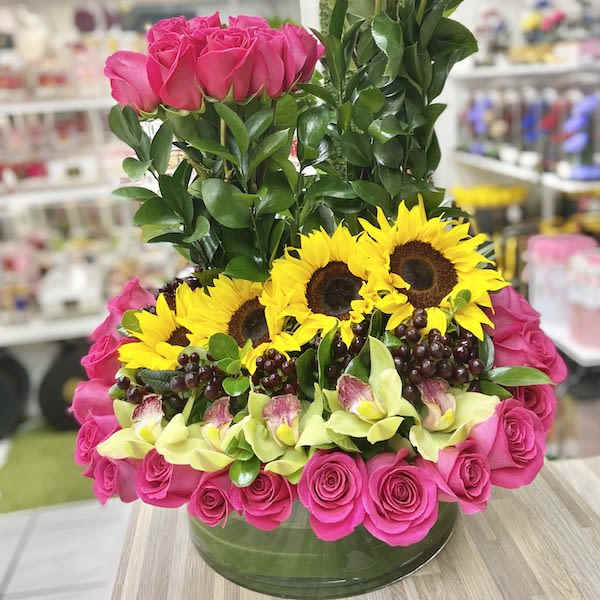 Structured arrangement of hot pink roses, yellow sunflowers, green orchids and grapes in a low glass vase
