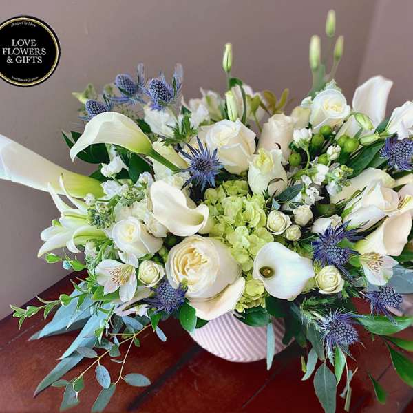 Low white and green arrangement of roses, calla lilies, and blue thistle in a white ribbed vase