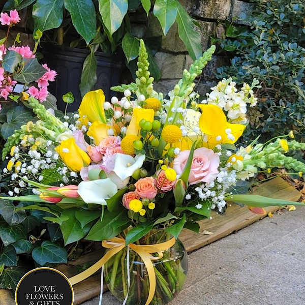 Bright bouquet of yellow calla lilies, pink roses, and white blooms in a clear glass vase with a yellow ribbon.