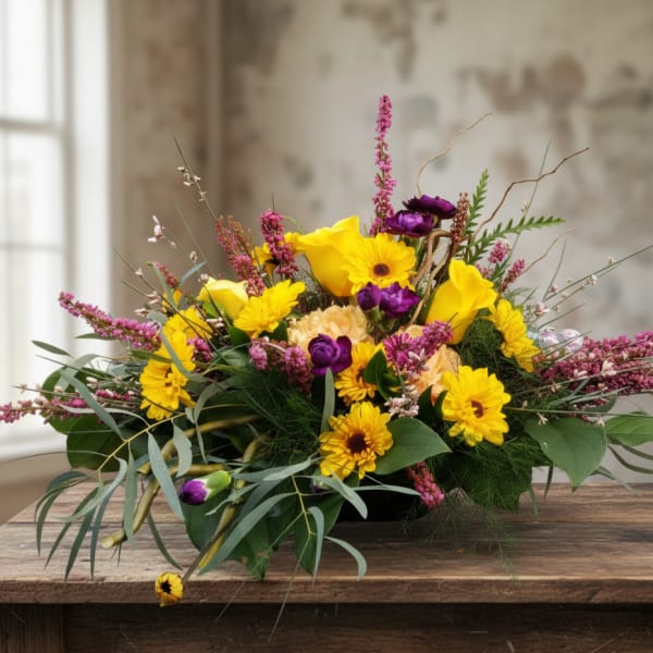 Low arrangement of yellow gerbera and calla lilies with purple blooms and pink spikes in a shallow container