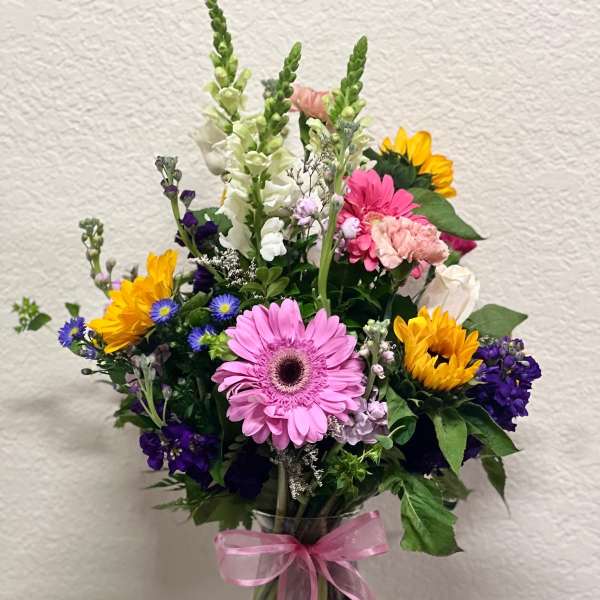 Mixed bouquet of pink, yellow, white, and purple flowers in a glass vase with a pink ribbon