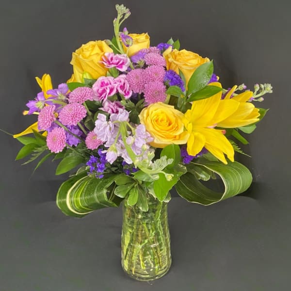 Yellow roses and purple flowers arranged in a glass vase.