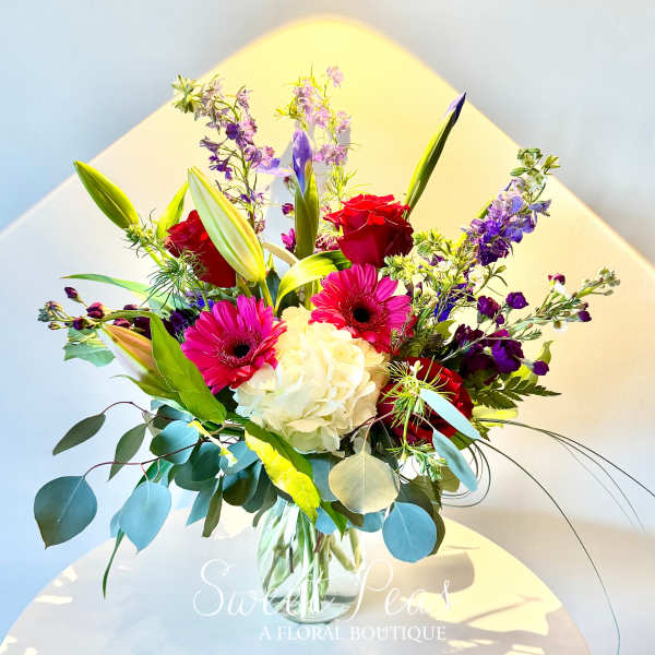 Bouquet of red roses, pink gerbera daisies, white hydrangea, and purple blooms in a glass vase