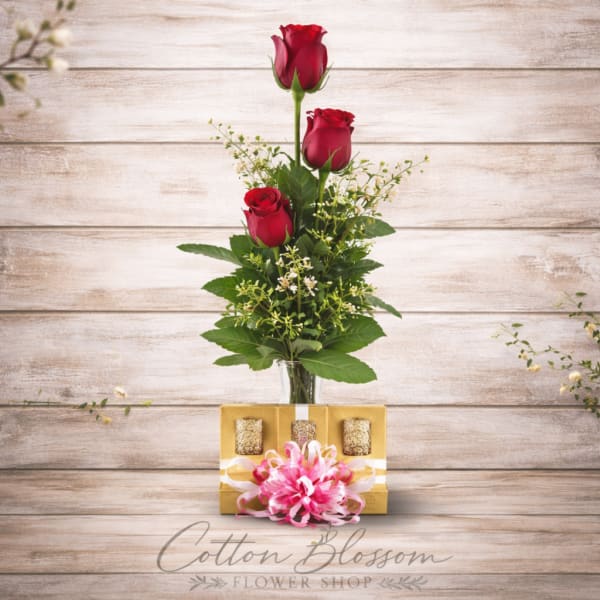 Three red roses in a glass vase with a gift box and pink bow