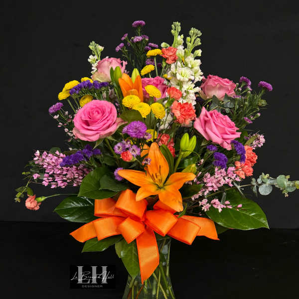 Mixed bouquet of pink, orange, yellow, and purple flowers in a glass vase with an orange ribbon
