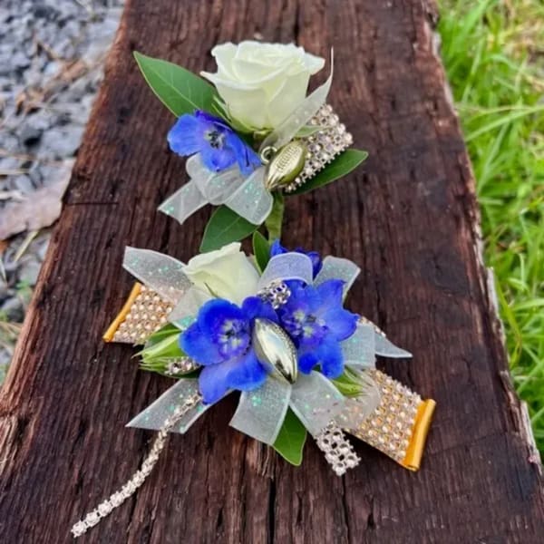 Two floral corsages with white roses and blue flowers on a wooden surface
