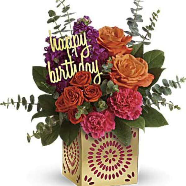 Birthday arrangement with orange roses, pink carnations, and purple flowers in a patterned cube vase.