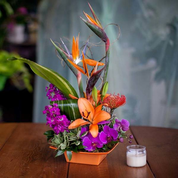 Tropical flower arrangement with orange lilies and purple orchids in an orange container