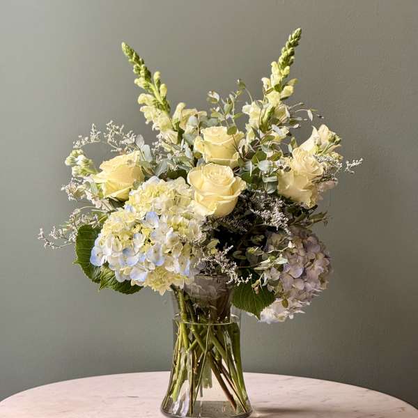 Arrangement of pale yellow roses, white hydrangeas, and snapdragons in a clear glass vase.