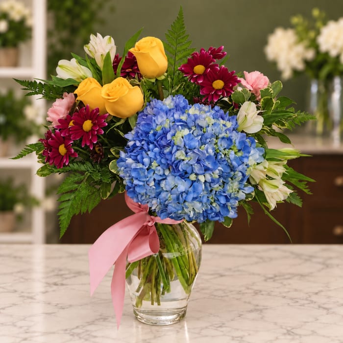 Mixed bouquet with blue hydrangea, yellow roses, and magenta daisies in a glass vase