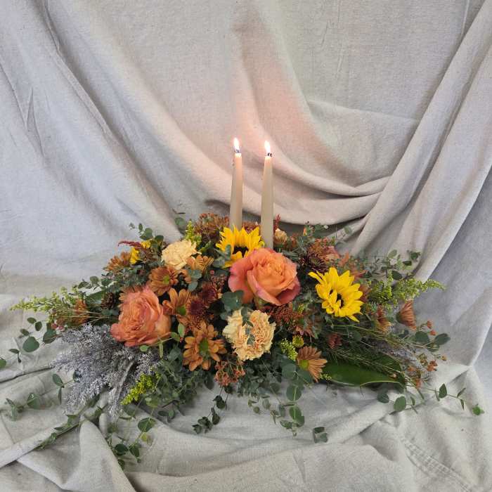 Low autumn centerpiece of orange roses, yellow sunflowers, and two lit taper candles on a neutral cloth backdrop.