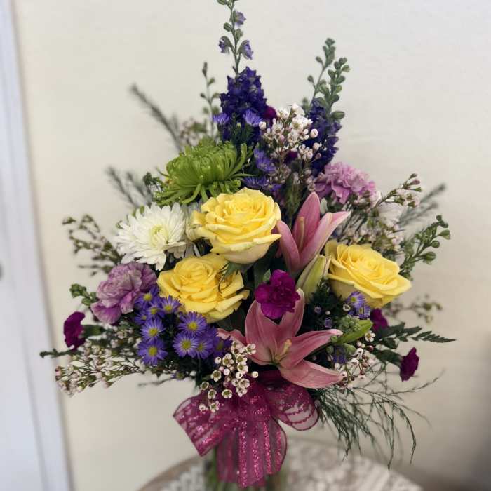 Mixed bouquet of yellow roses, pink lilies, and purple flowers in a glass vase