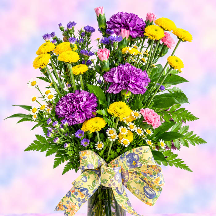 Mixed bouquet of purple, yellow, and pink flowers with a patterned bow in a vase
