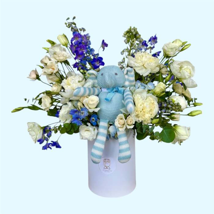 Blue teddy bear in a white box surrounded by white and blue flowers
