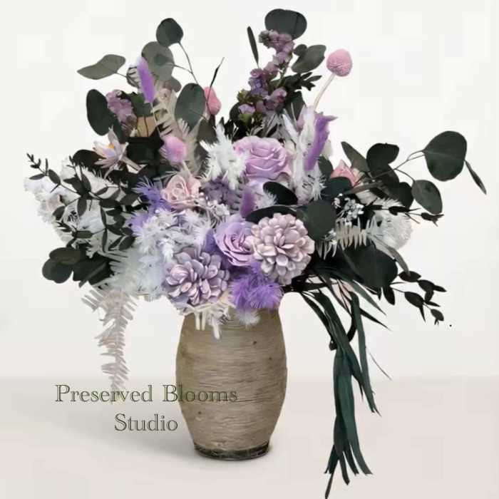 Tall arrangement of lavender and white preserved flowers with dark greenery in a textured beige vase