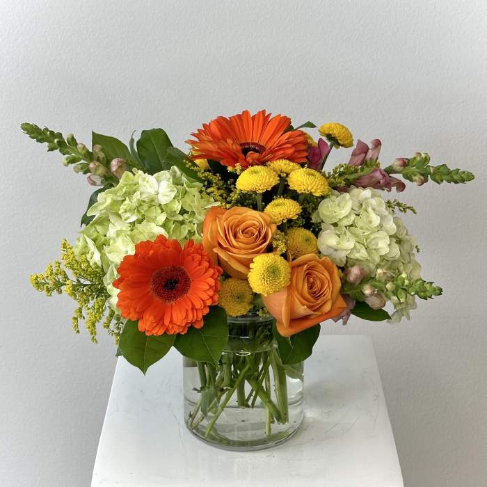 Orange gerbera daisies and roses in a clear glass vase with pale hydrangeas
