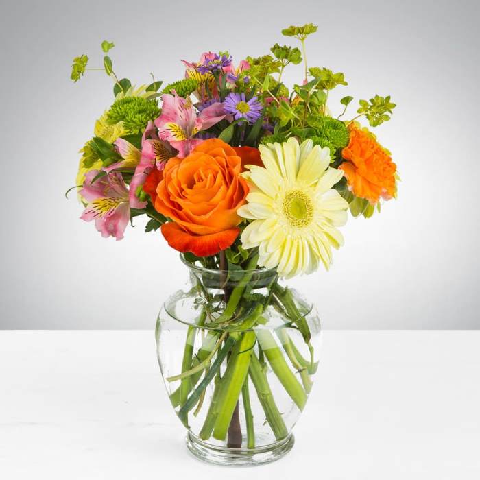 Bright mixed bouquet of orange roses, yellow gerbera daisy, and other blooms in a clear glass vase