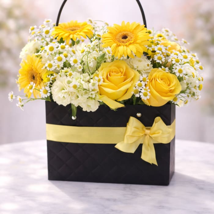Yellow roses and gerbera daisies with white daisies in a black gift bag with yellow ribbon.