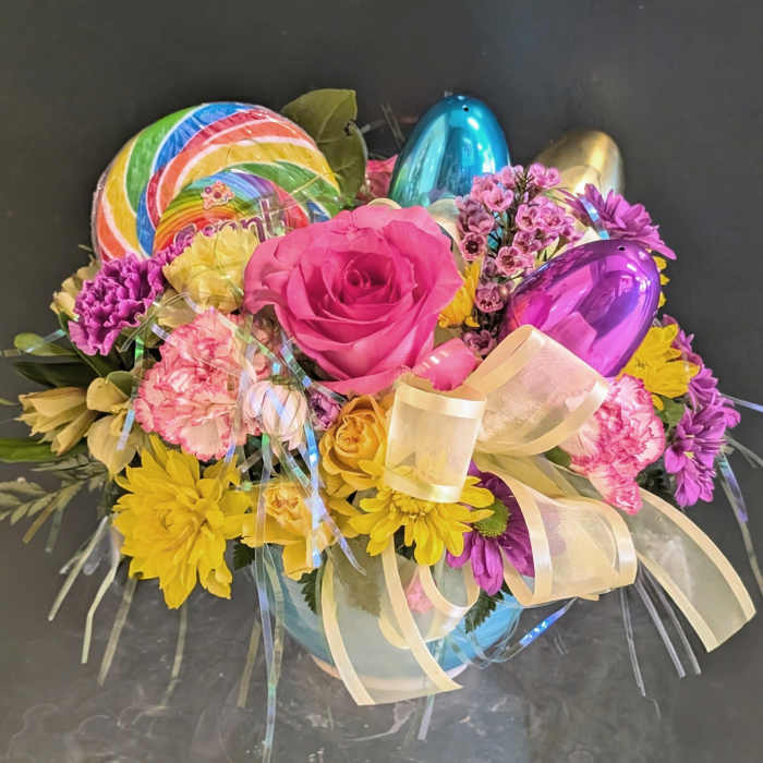 Colorful spring flower arrangement with pink rose, yellow daisies, lollipop, ribbon, and metallic Easter eggs in a bowl vase