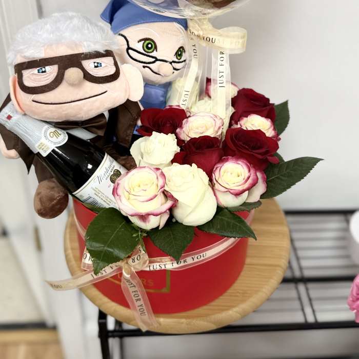 Gift set with red and white roses in a red box, two plush dolls, a bottle, and a clear anniversary balloon.