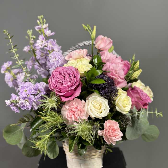 Basket arrangement of pink and cream roses with lavender stock and green thistle blooms