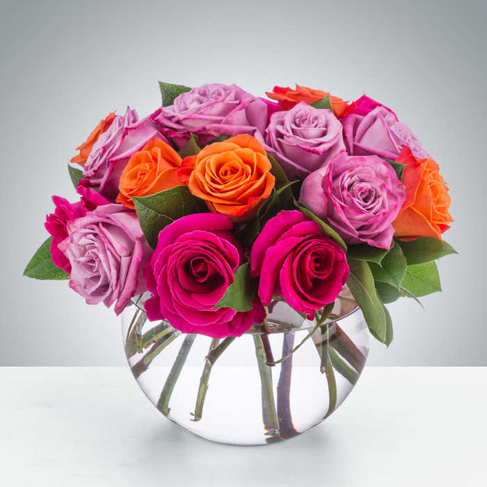 Compact arrangement of pink, lavender, and orange roses in a clear round glass vase