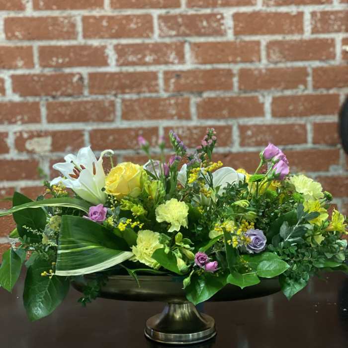Low floral centerpiece with yellow roses, white lilies, and pink blooms in a pedestal bowl.