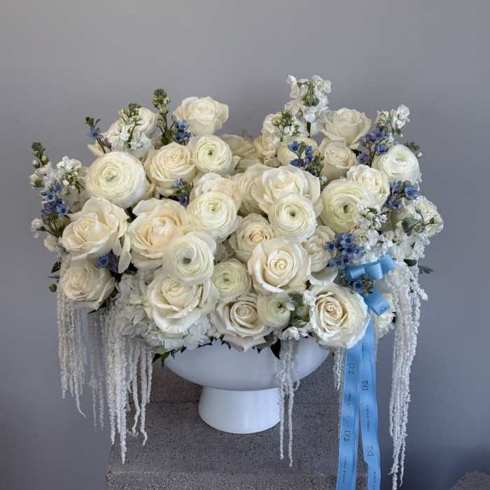 White rose and ranunculus arrangement in a white pedestal vase with blue ribbon