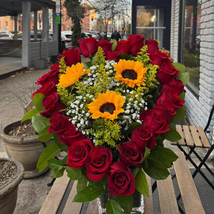 Heart-shaped arrangement of red roses with yellow sunflowers and white filler flowers in a clear glass vase
