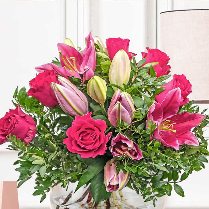 Arrangement of hot pink roses and pink lilies in a round clear glass vase on a wooden shelf