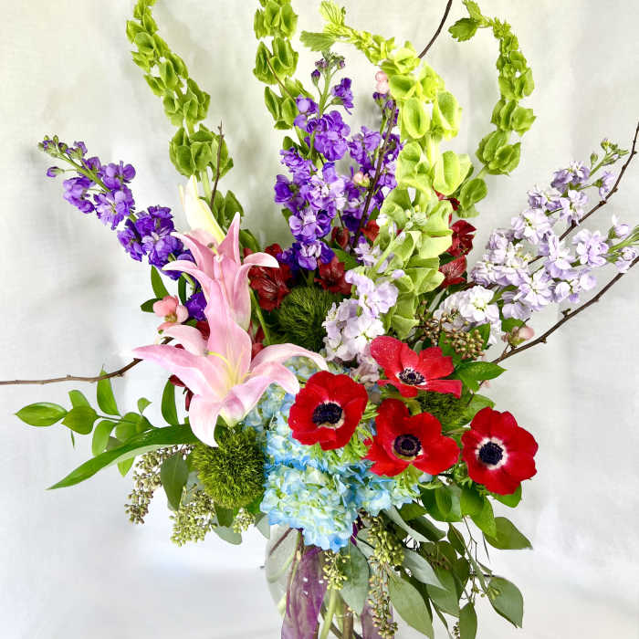 Tall bouquet of pink lilies, red anemones, and purple blooms in a glass vase