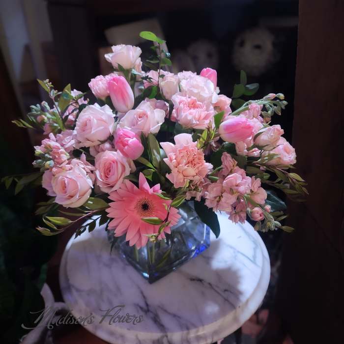 Low arrangement of pink roses, tulips, carnations, and a gerbera daisy in a glass cube vase.