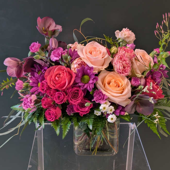 Low rectangular bouquet of pink, peach, and purple flowers in a clear glass vase