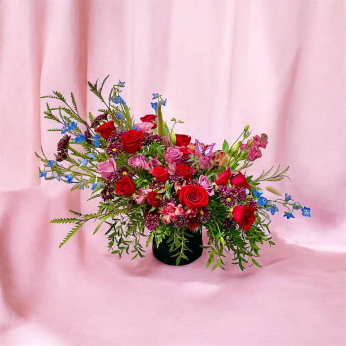 Asymmetrical arrangement of red and pink roses with blue accents in a black vase on a pink backdrop