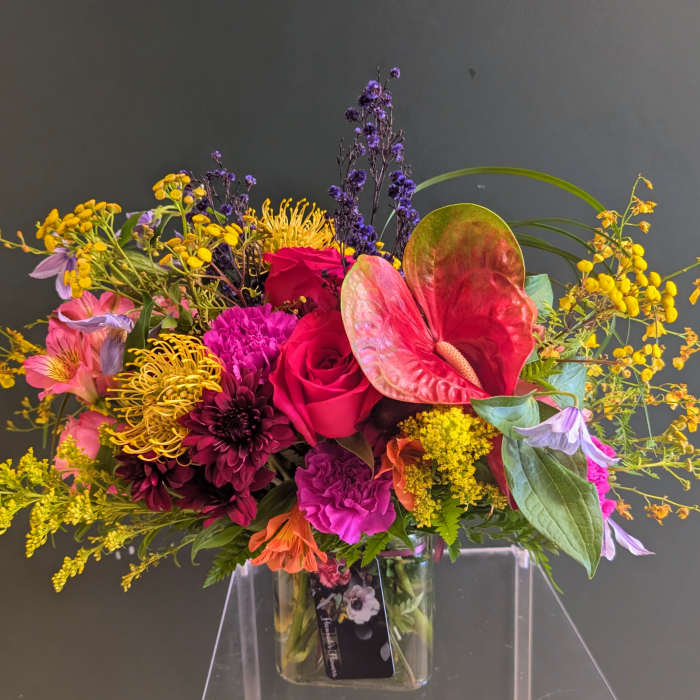 Bright mixed arrangement with pink roses, yellow pincushion blooms, and a red tropical flower in a glass cube vase
