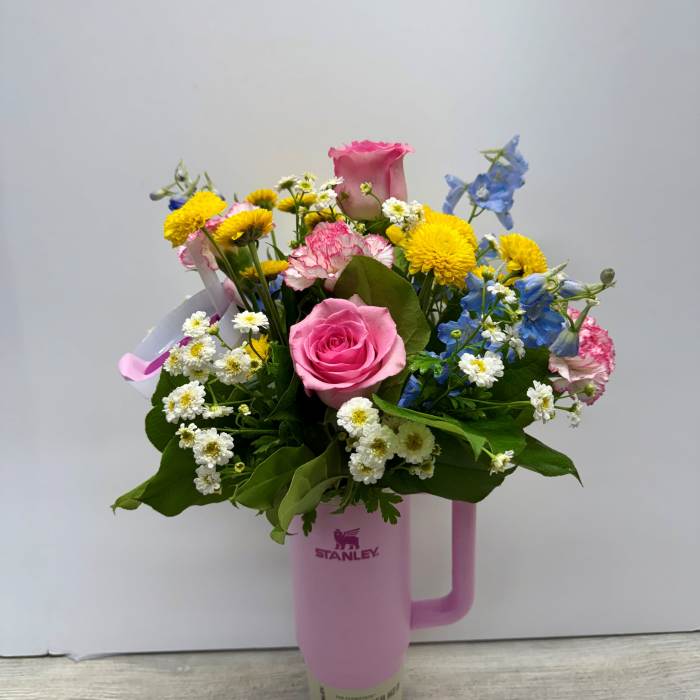 Mixed bouquet of pink roses, carnations, yellow mums, and blue blooms arranged in a pink tumbler.