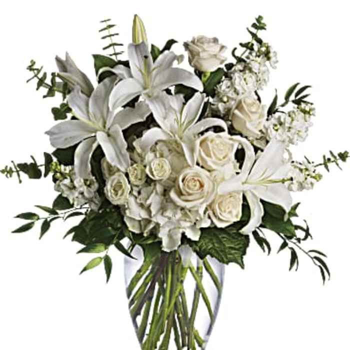 Tall arrangement of white lilies, roses, and other white flowers in a clear glass vase