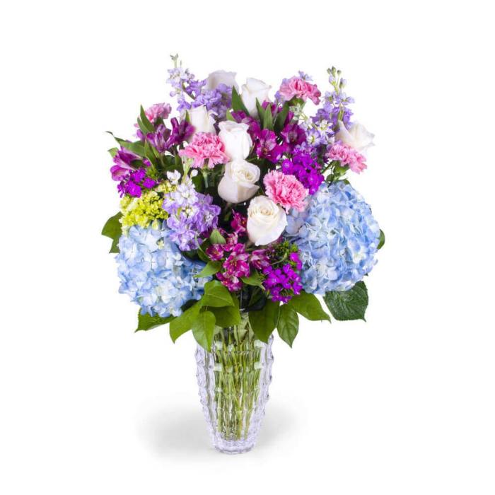 Tall mixed bouquet of white roses, blue hydrangeas, and pink and purple blooms in a clear glass vase