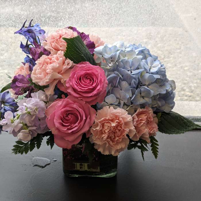 Low arrangement of pink roses, peach carnations and blue hydrangeas in a clear glass cube vase