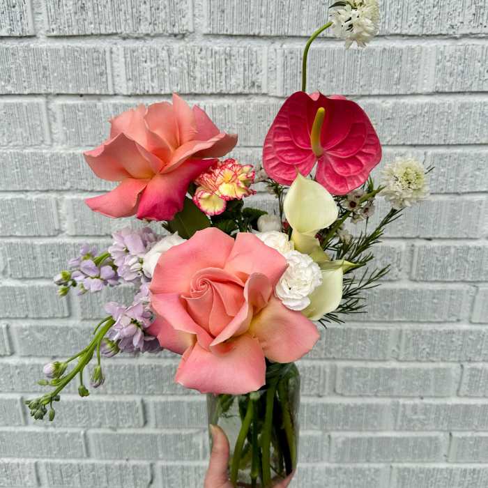 Tall mixed arrangement of peach roses, pink anthurium, white calla lilies, and other blooms in a clear glass vase.