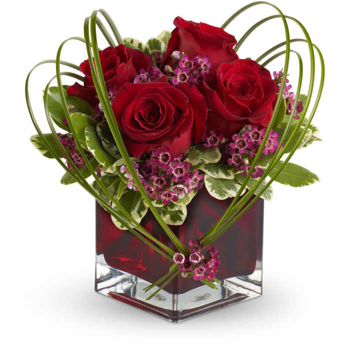 Red rose arrangement with pink blossoms in a glass cube vase framed by heart-shaped loops of grass.