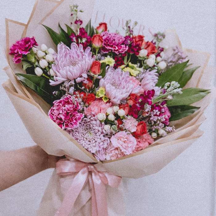 Handheld bouquet of pink and red flowers wrapped in beige paper with a pink ribbon