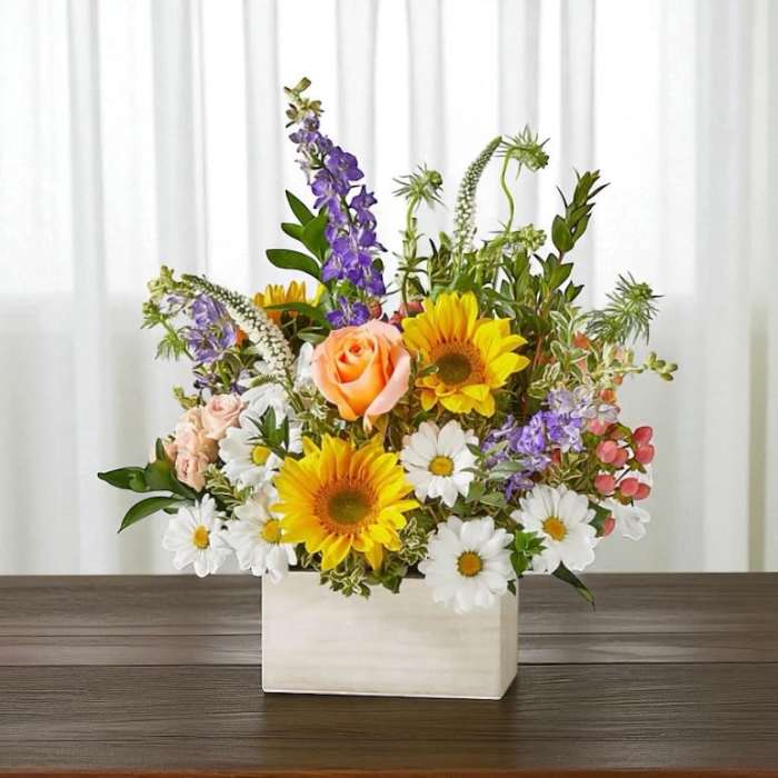 Mixed bouquet in a white rectangular vase with sunflowers, roses, and daisies
