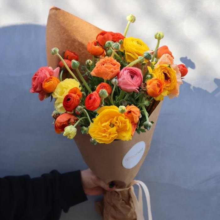 Handheld bouquet of orange, yellow, and pink ranunculus wrapped in brown paper