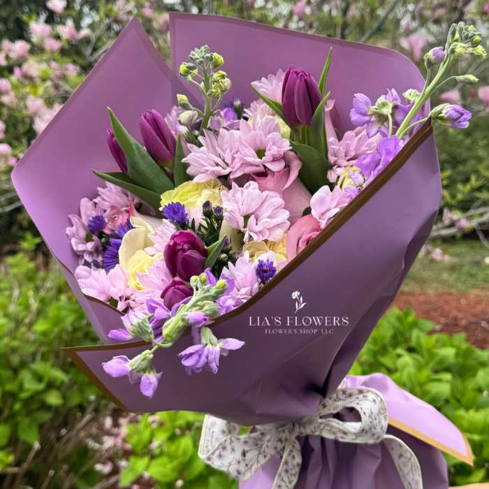 Purple-wrapped bouquet of pink, lavender, and yellow flowers with a ribbon tie