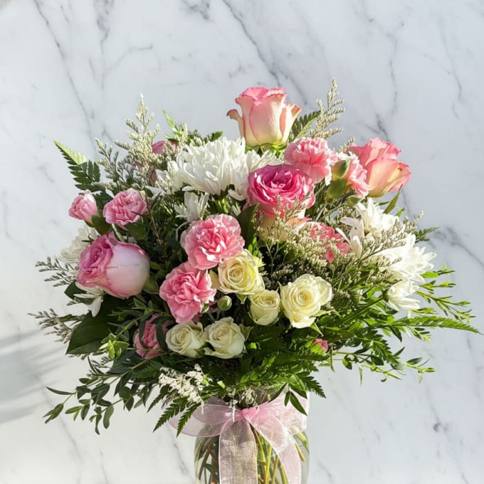 Pink and white bouquet of roses, carnations, and daisies in a glass vase with a ribbon.