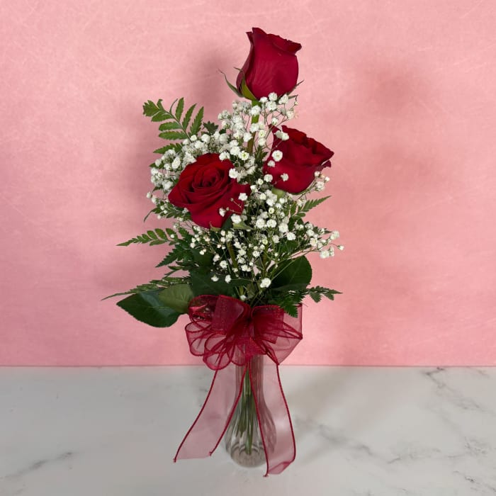 Three red roses in a clear vase with baby's breath and a red ribbon