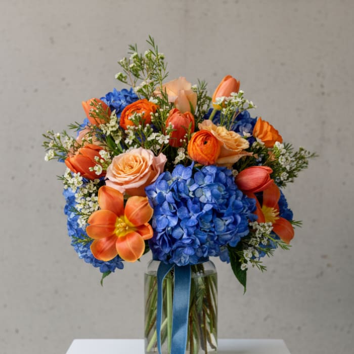 Bouquet of blue hydrangeas, orange tulips, and peach roses in a glass vase